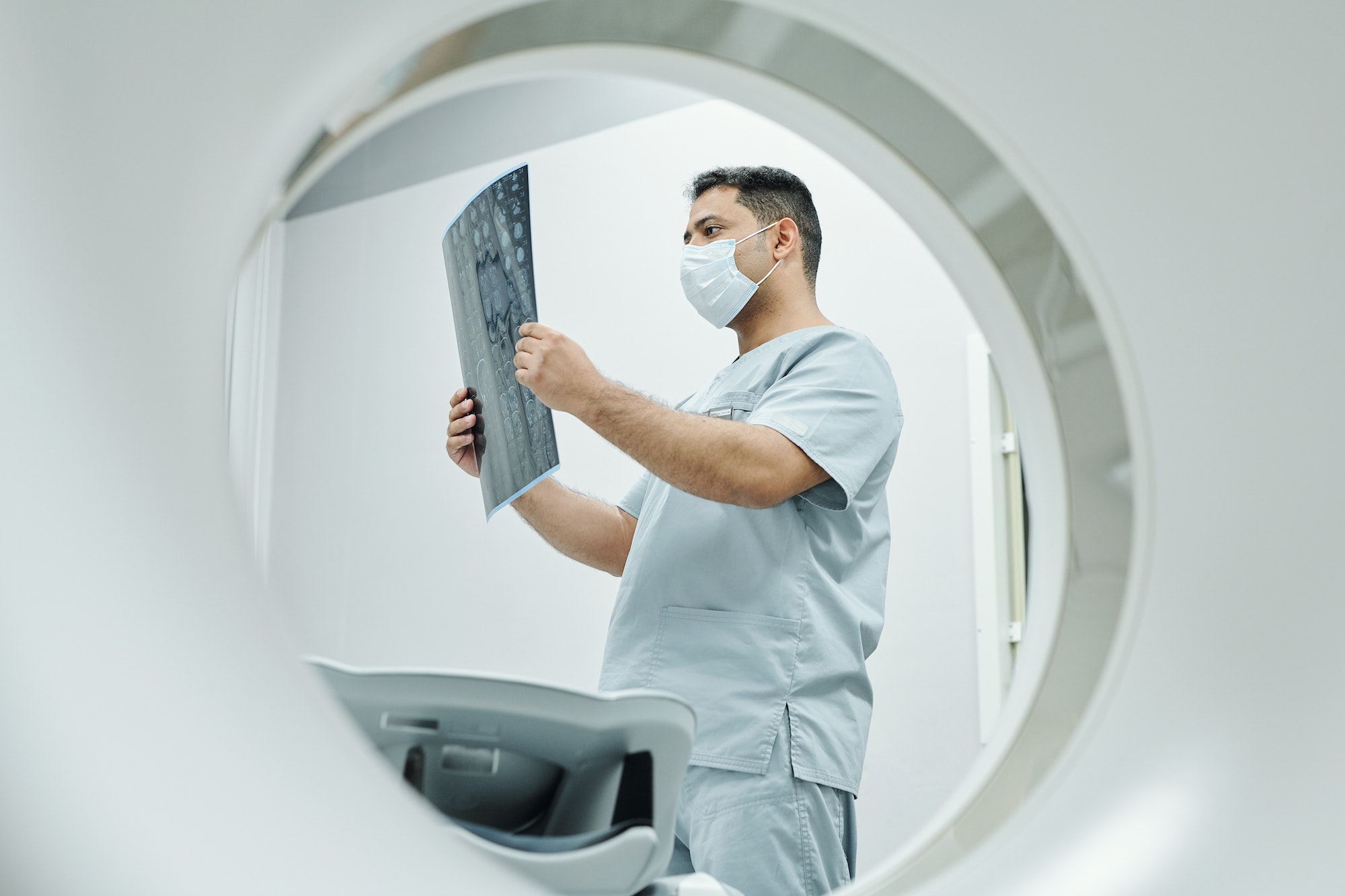 Serious mature mixed-race radiologist in mask and uniform looking at x-ray image